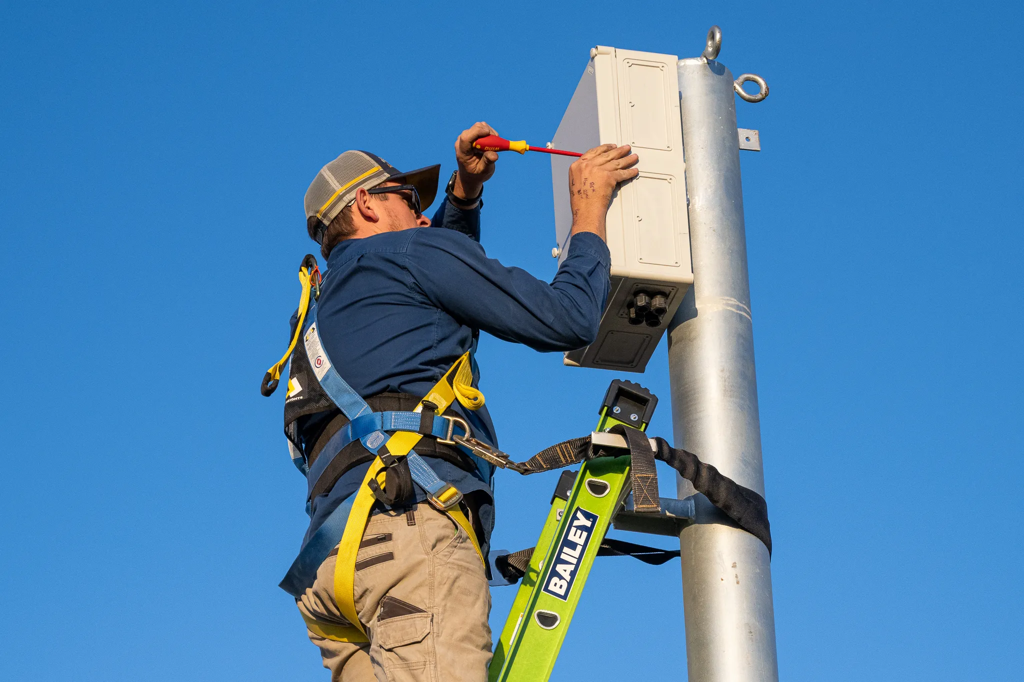 A Switch'D electrician harnessed on a ladder fitting a pole-top enclosure against a clear sky